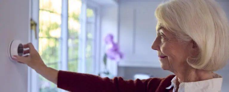 Senior Woman At Home Turning Down Central Heating Thermostat During Cost Of Living Energy Crisis